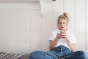 Blondes Teenie-Mädchen mit wiessem T-Shirt und Jeanshose sitzt auf dem Bett, an die weisse Holzwand gelehnt und hält mit beiden Händen sein Smartphone, in das es konzentriert blickt Blondes Teenie-Mädchen mit wiessem T-Shirt und Jeanshose sitzt auf dem Bett, an die weisse Holzwand gelehnt und hält mit beiden Händen sein Smartphone, in das es konzentriert blickt